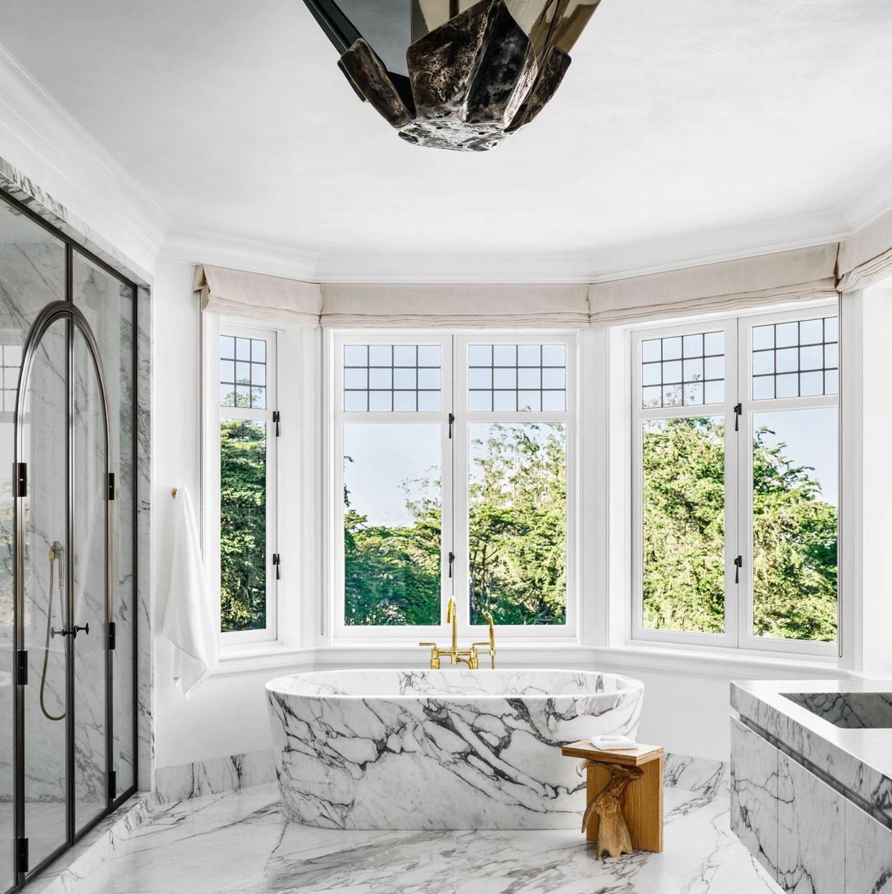 rounded bathroom with double windows in an arc at the back with a standalone marble tub at center and a floating marble vanity and a large bat like light fixture at center and a glass shower at left