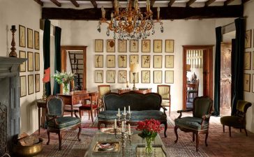 a room with a crystal chandelier, vintage sofa and chairs with velvet cushions, cocktail table with candelabras and trays, oriental rug, wooden desk and chair, a wall with 21 small framed botanical art pieces, beamed ceiling