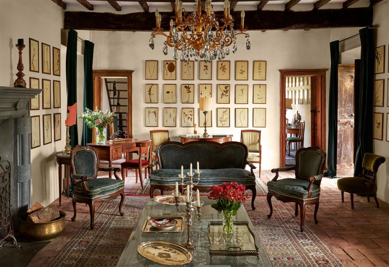 a room with a crystal chandelier, vintage sofa and chairs with velvet cushions, cocktail table with candelabras and trays, oriental rug, wooden desk and chair, a wall with 21 small framed botanical art pieces, beamed ceiling