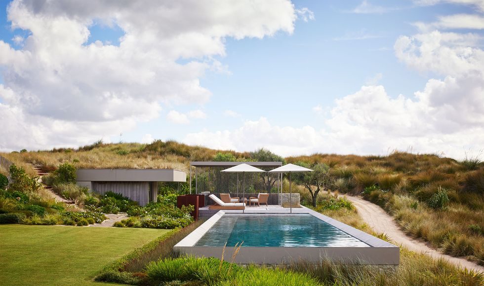 by a dirt road is a swimming pool with umbrellas and chairs by a pool house built into the grassy hillside