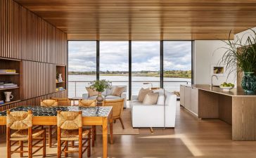 a large open room with a glass wall looking out to the sea, dining table and chairs, sofa, loveseat, and cocktail table, slatted wood wall with inset shelves, kitchen island with sink and cabinets, wood ceiling and floor