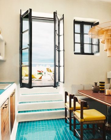 a kitchen with turquoise tiled floor and a wood table with yellow chairs and a banquette with striped fabric, rattan pendant, counter with sink and shelves above and wood cabinets below, steps go to a doorway out to terrace