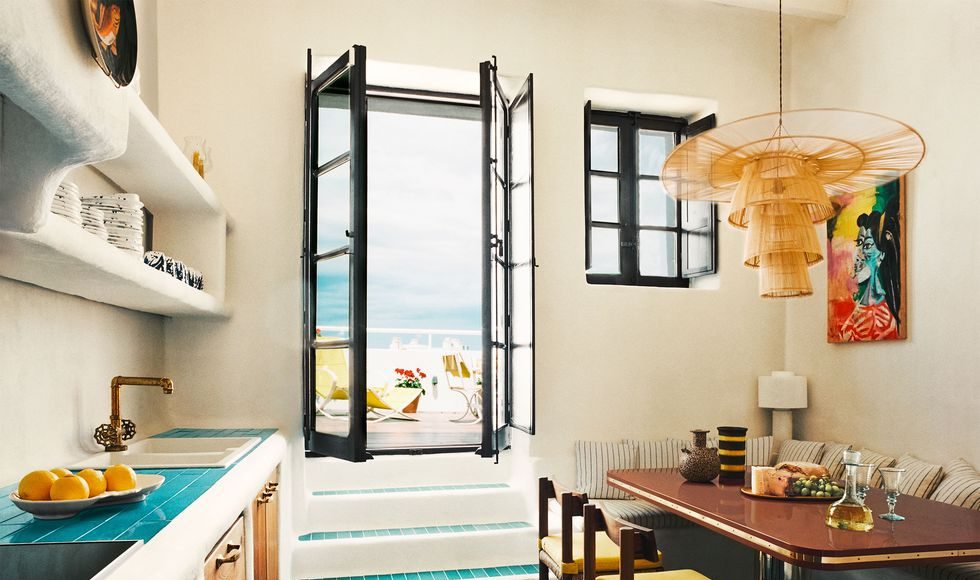 a kitchen with turquoise tiled floor and a wood table with yellow chairs and a banquette with striped fabric, rattan pendant, counter with sink and shelves above and wood cabinets below, steps go to a doorway out to terrace