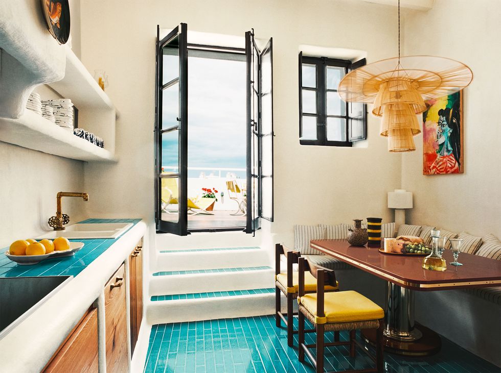 a kitchen with turquoise tiled floor and a wood table with yellow chairs and a banquette with striped fabric, rattan pendant, counter with sink and shelves above and wood cabinets below, steps go to a doorway out to terrace