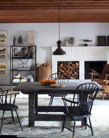 wood beam ceiling living dining space with a white brick fireplace at far end and a wood insert next to it and some modern wood club chairs and a dark square farmhouse table and chairs in the foreground and a kitchen set up to the left