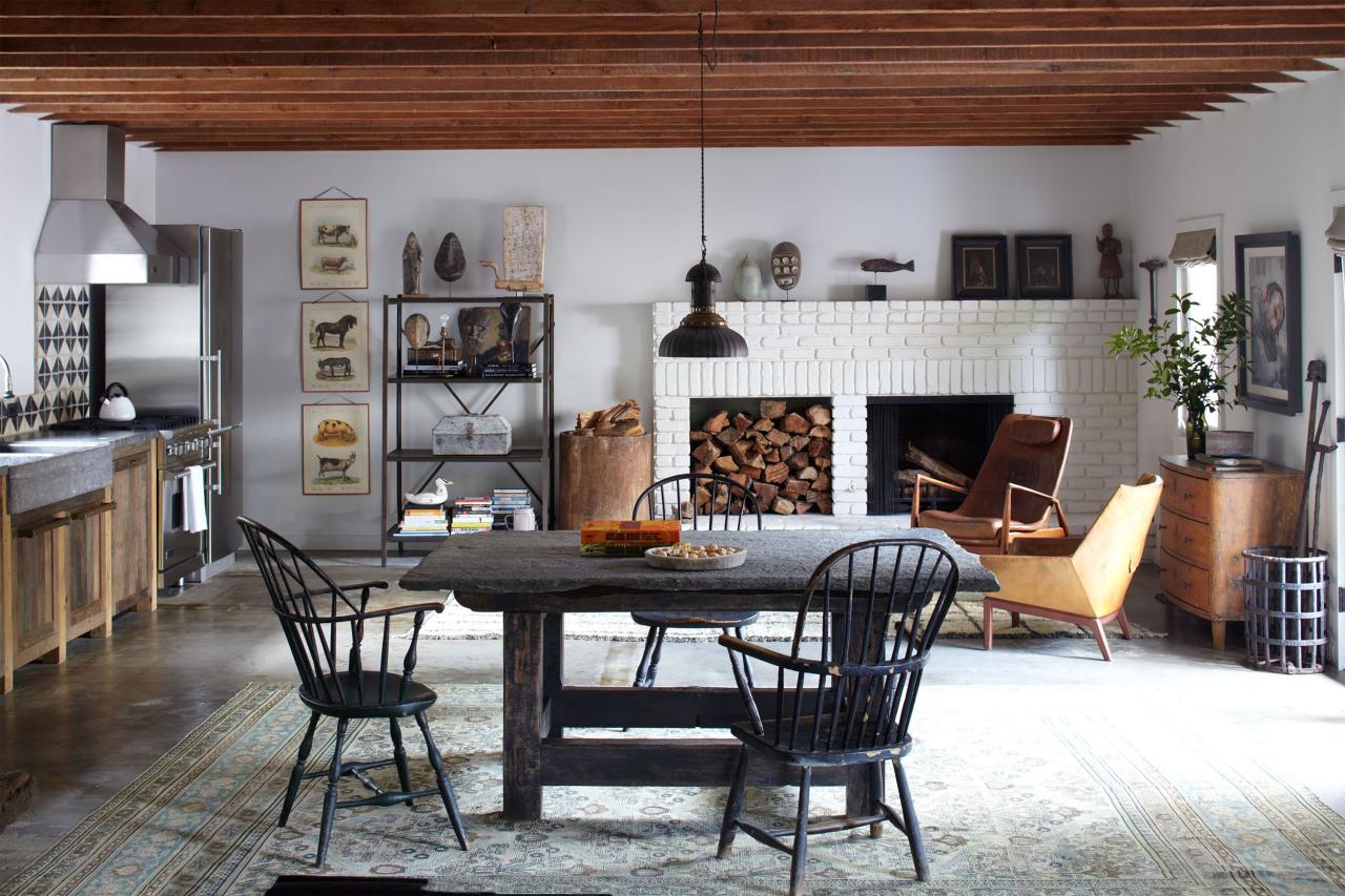 wood beam ceiling living dining space with a white brick fireplace at far end and a wood insert next to it and some modern wood club chairs and a dark square farmhouse table and chairs in the foreground and a kitchen set up to the left