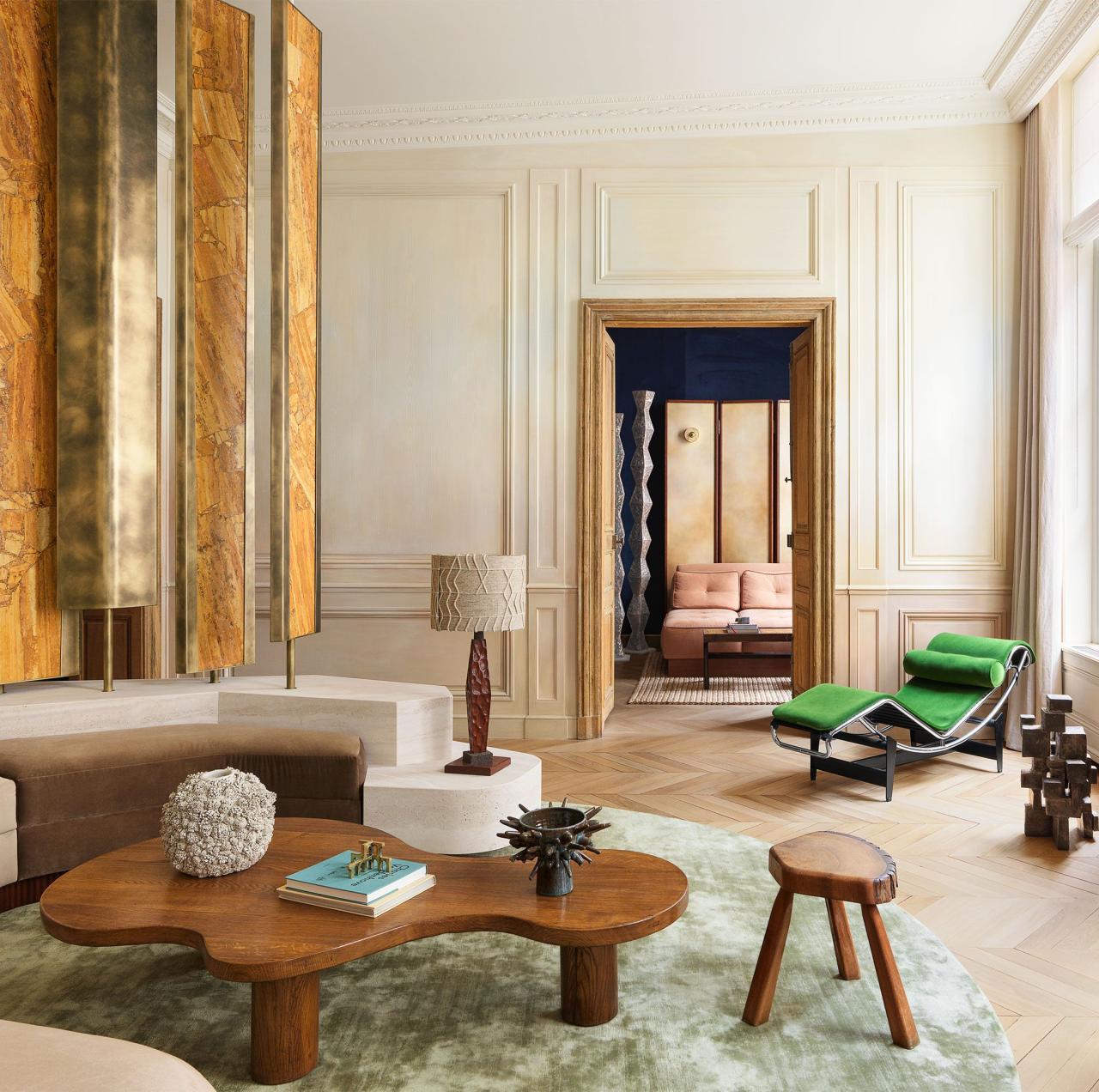 living room with beige armless sofa and four decorative rotating yellow travertine panels on a platform behind it, a curvy wooden cocktail table, a vintage lamp, stool, a bright green chaise, and a muted green rug